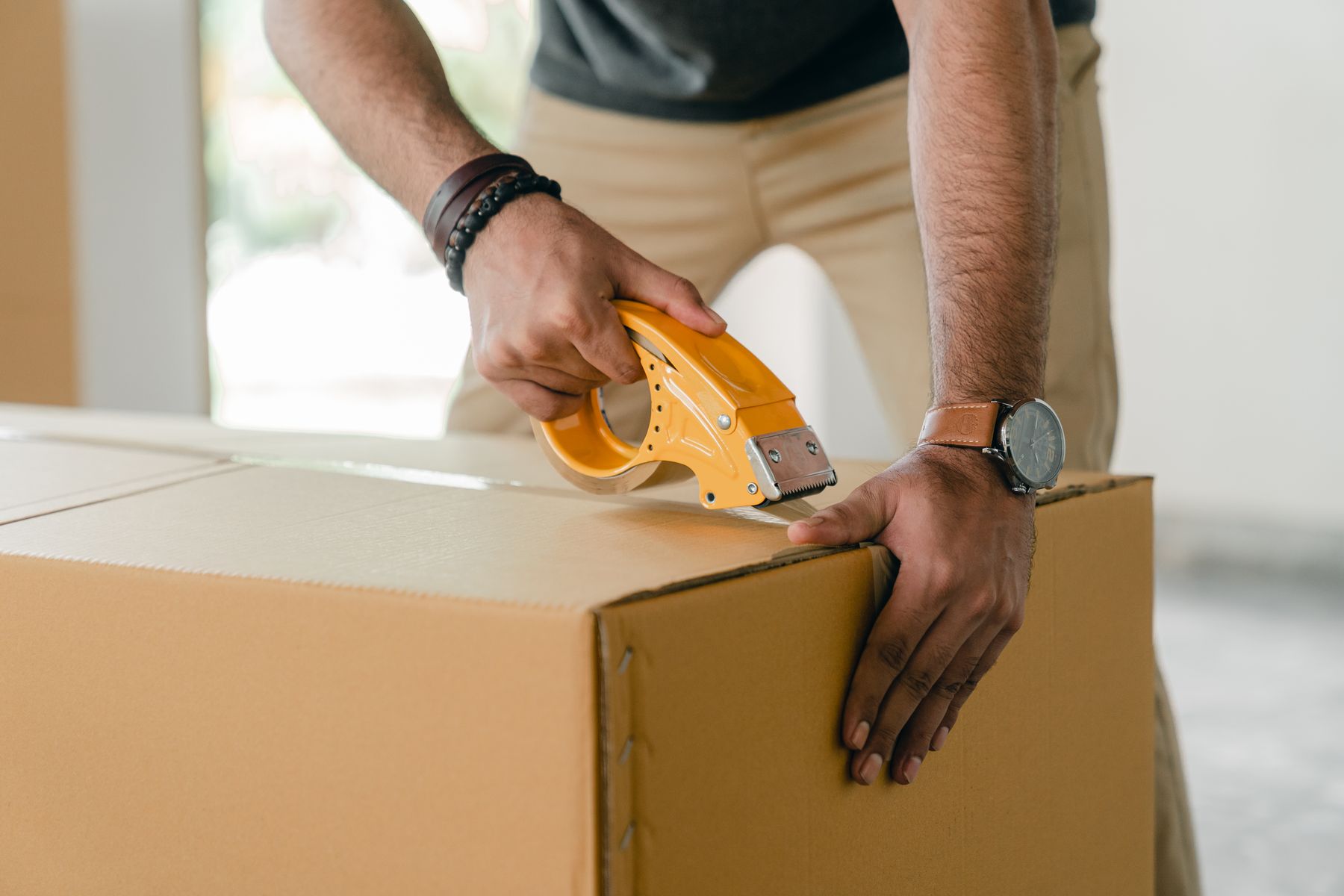 Hands sealing a plain brown carton with a tape dispenser.